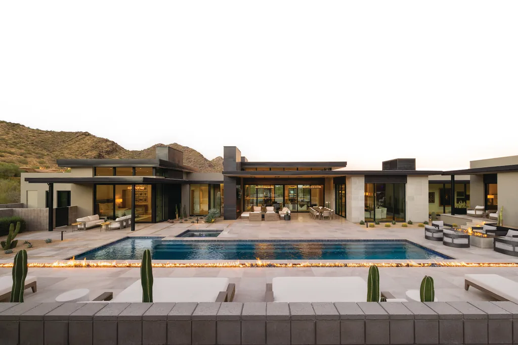 A modern single-storey desert home at dusk, its glass walls lit, a long rectangular pool and lounge deck in front, cactus and desert planting along the pool edge and mountains behind.
