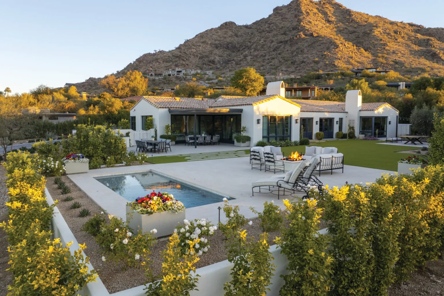 A hillside home with a pool and spa on a stone terrace at golden hour, yellow flowering shrubs in the foreground and a rocky mountain behind.