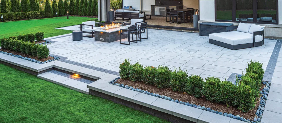 A large light stone patio with a sunken lounge and fire table, outlined by clipped hedges and set into a bright green lawn behind a modern home.