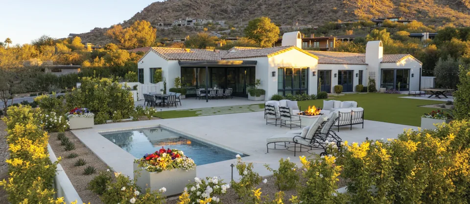 A pool and spa on a stone terrace of a hillside home at golden hour, with yellow flowering shrubs below and a rocky mountain behind.